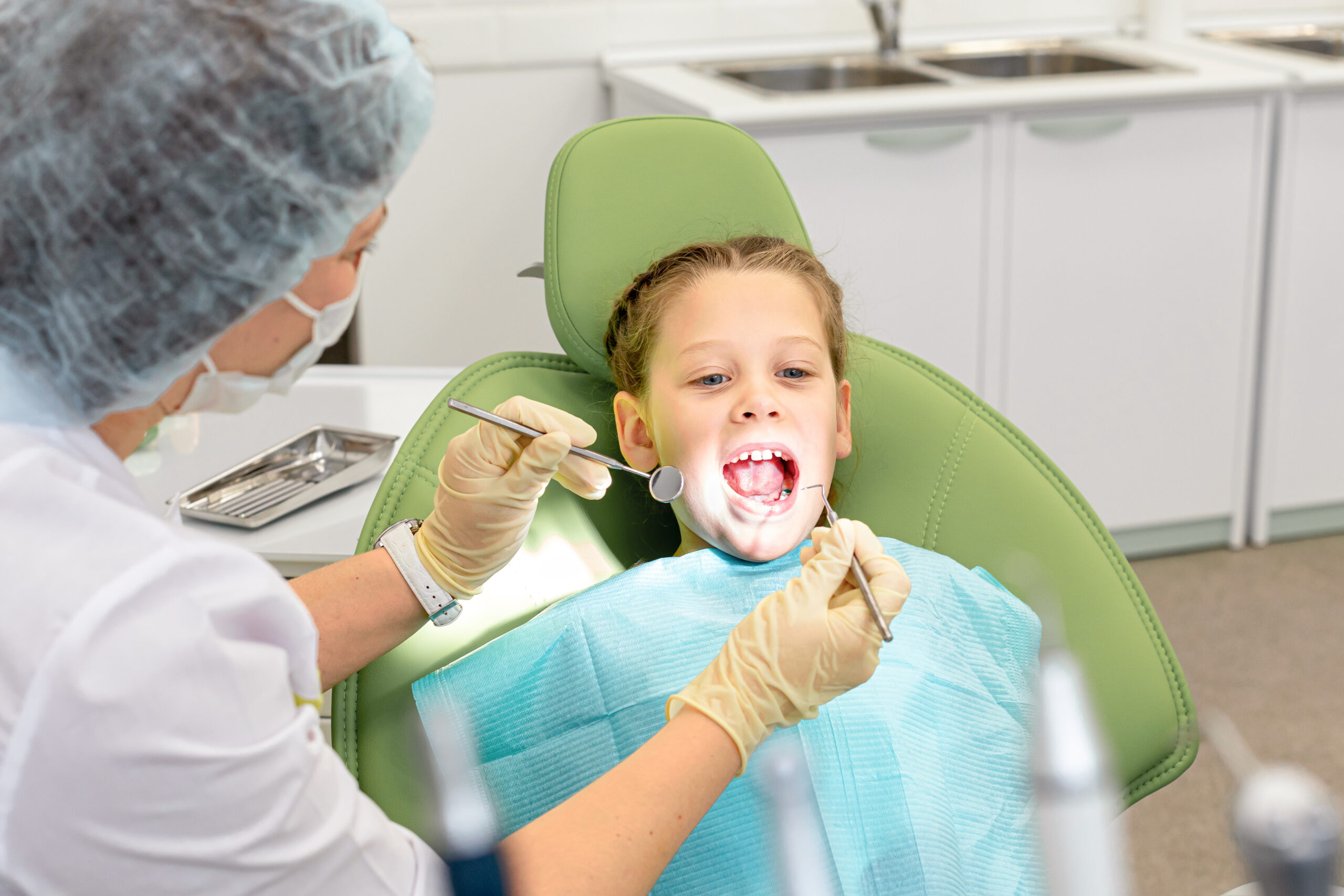 Little cute girl is sitting in dental chair and opening mouth in clinic, office. Doctor is preparing for examination of child teeth with instruments