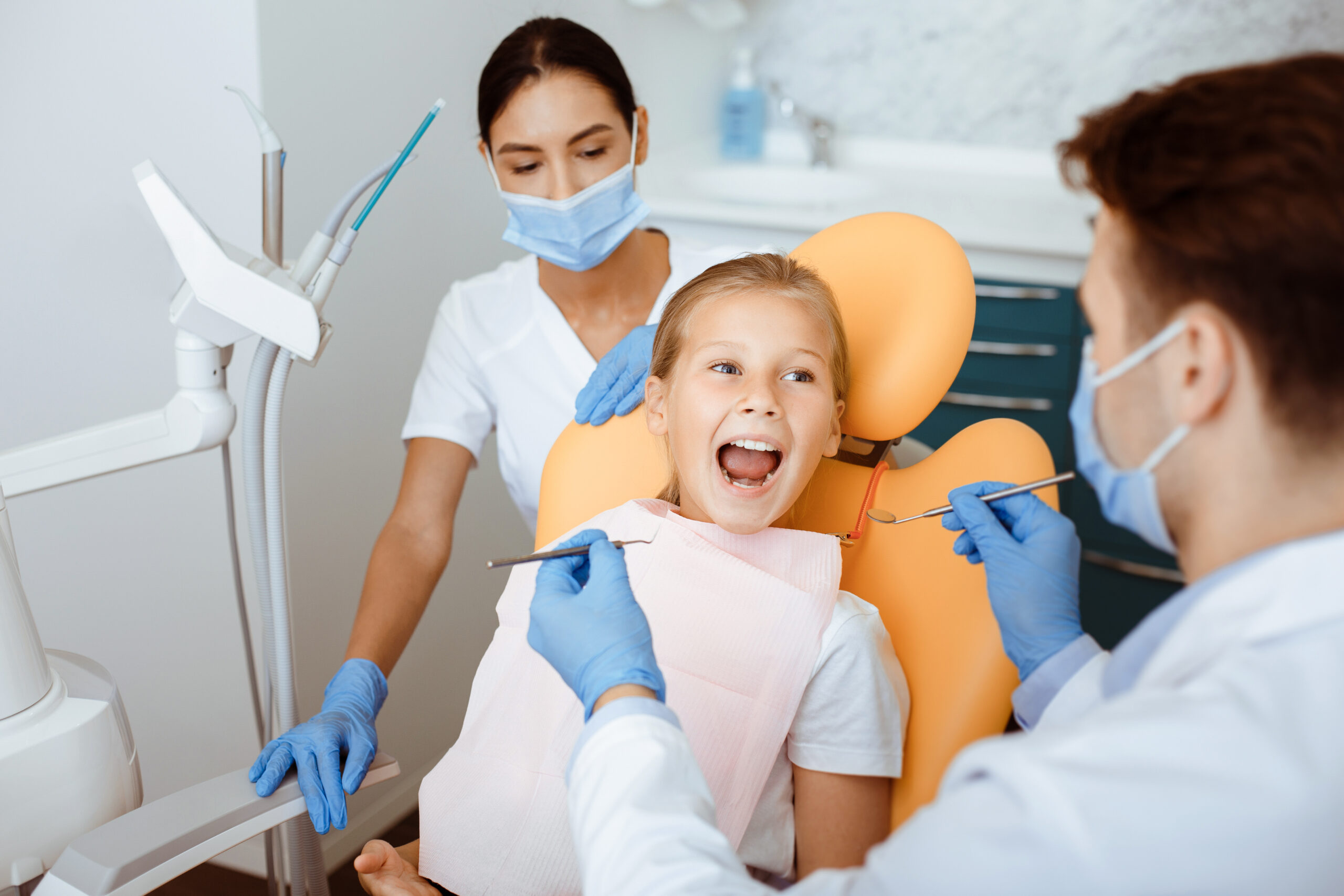 Work of professional doctor and pediatric dentist in modern clinic with equipment. Millennial man in white coat, protective mask and gloves with nurse treat teeth to little girl in chair, copy space