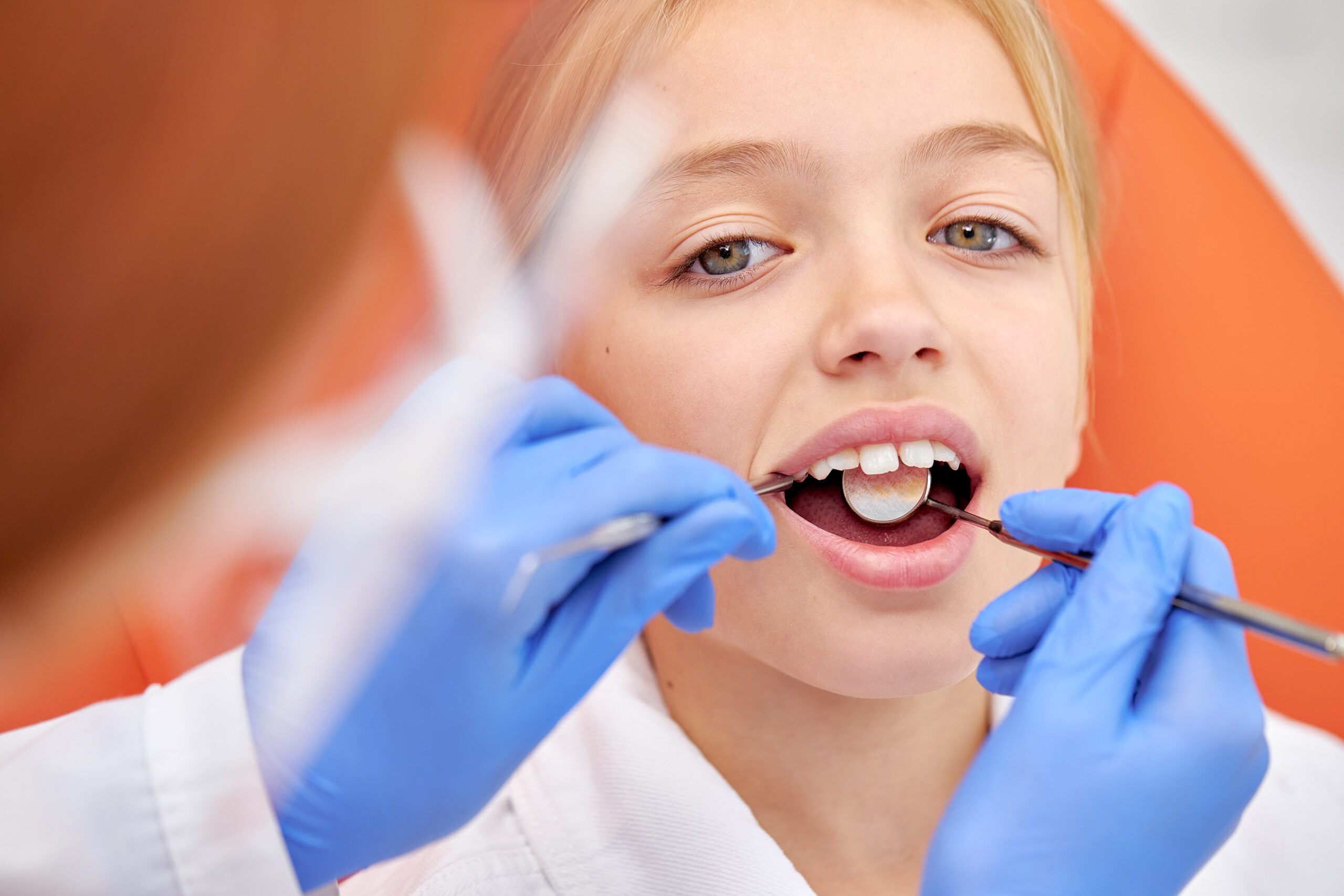 Close-up photo of child girl during dental tooth examination checkup. Attractive pretty caucasian kid sits with opened mouth while cropped female dentist in gloves examining inside of oral cavity