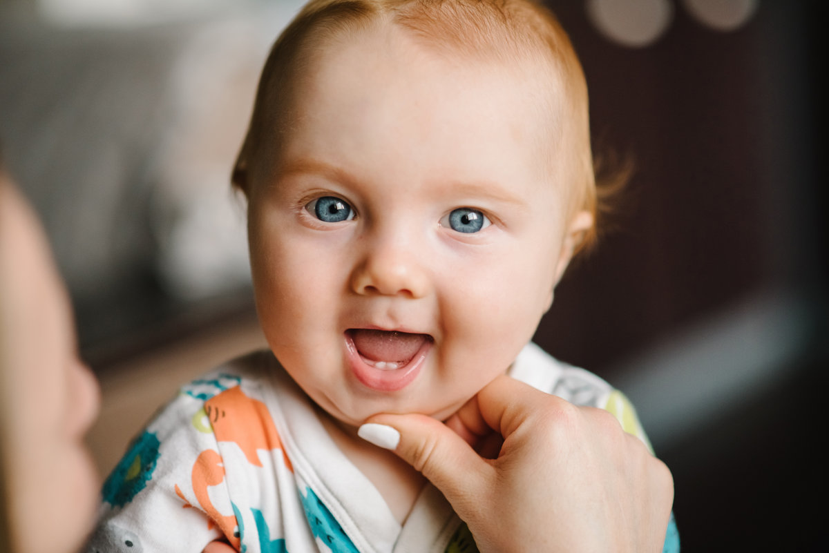 Baby boy portrait looking at camera. Close-up mother hands open baby mouth to examine first teeth. Infant primary tooth. The happy baby infant with first tooth.