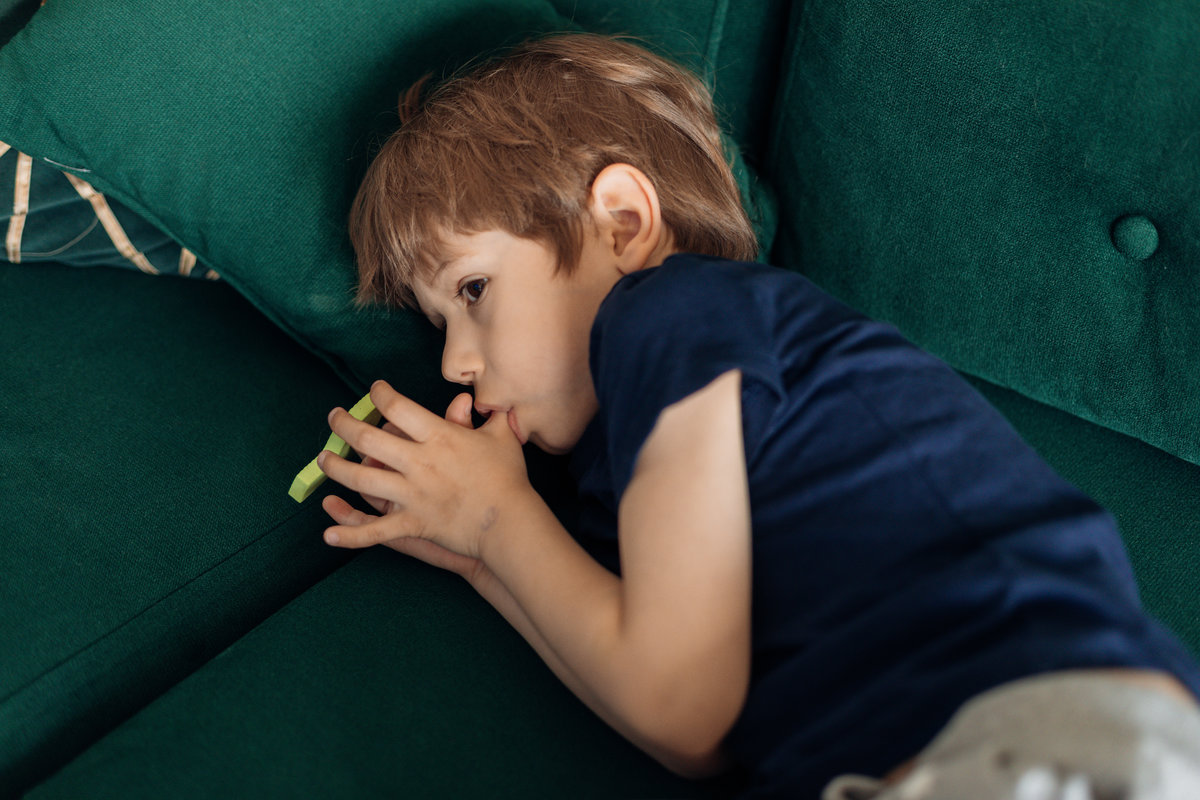 Portrait of serious, sad and depressed little boy lying on green sofa, sucking his thumb finger. Children stuttering, traumatic experience. Silent scary cry. Psychological stress and shock. Top view
