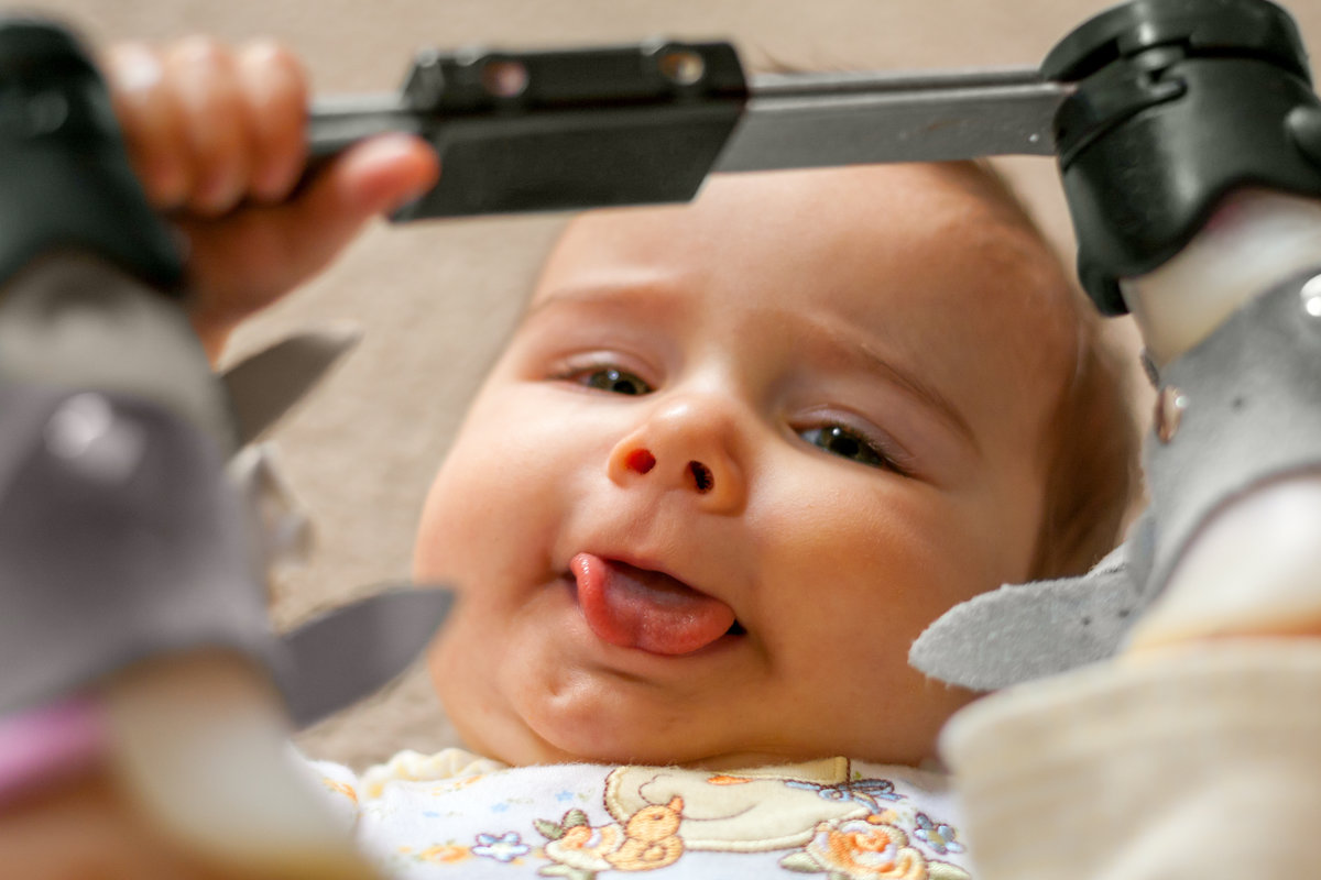 A nine month old baby with a tongue tie and orthopedic shoes lays on her back with her tongue sticking out as much as possible as she holds the bar between her orthopedic shoes that she wore for a club foot.