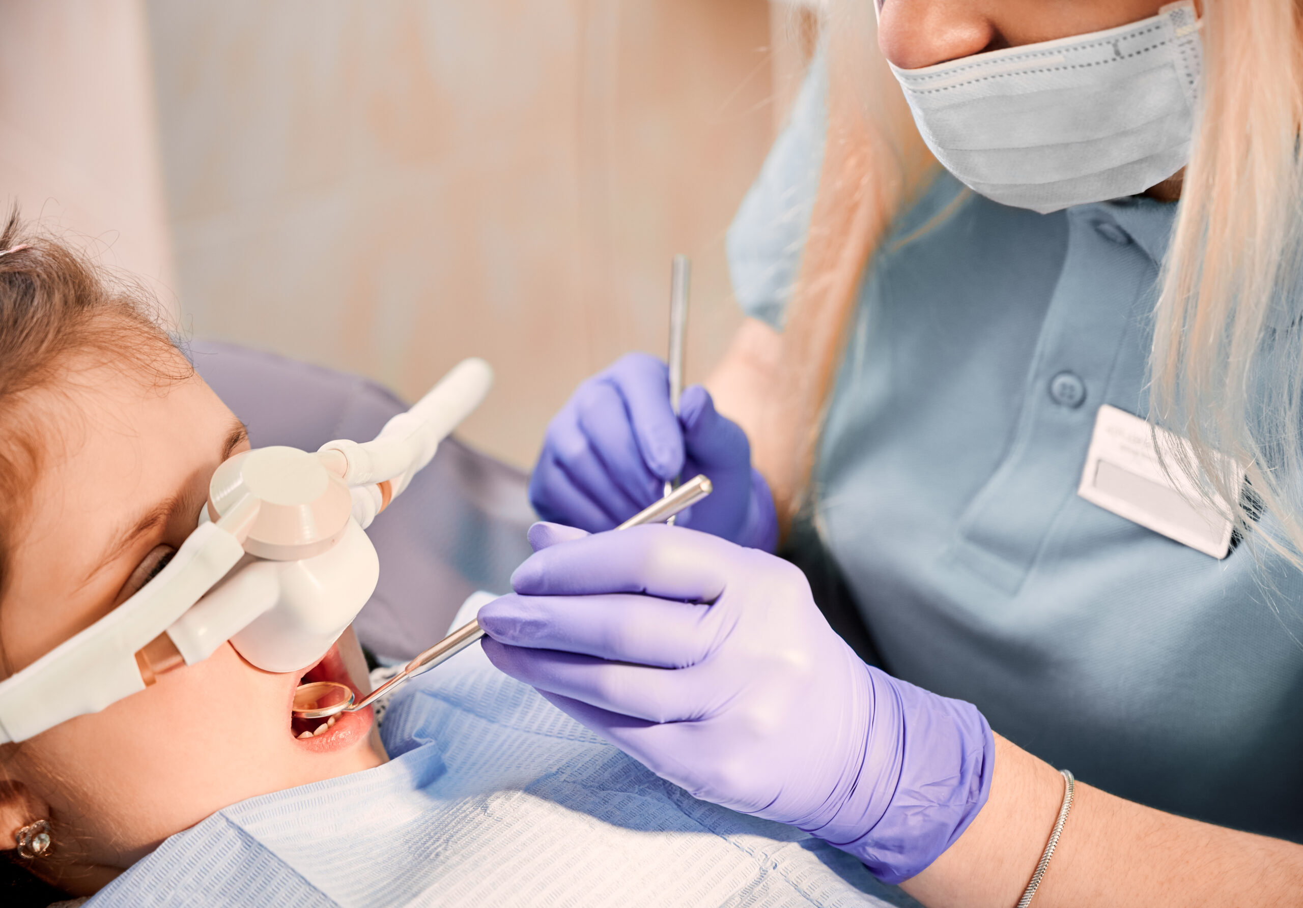 Female dentist checking child teeth with dental explorer and mirror while girl lying in dental chair with inhalation sedation at dental office. Concept of pediatric, sedation dentistry.