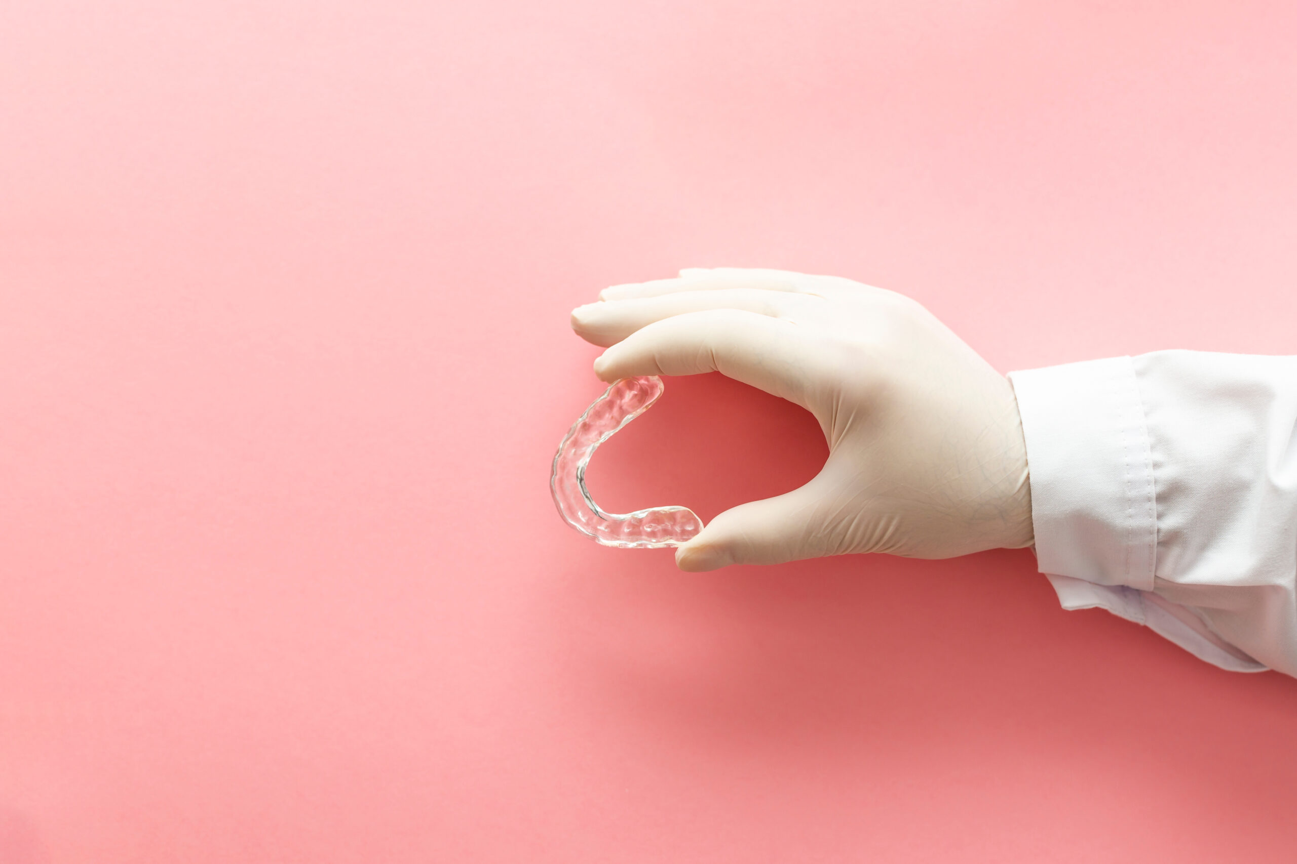 Hands of a dentist holding a transparent orthodontic with a pink background.